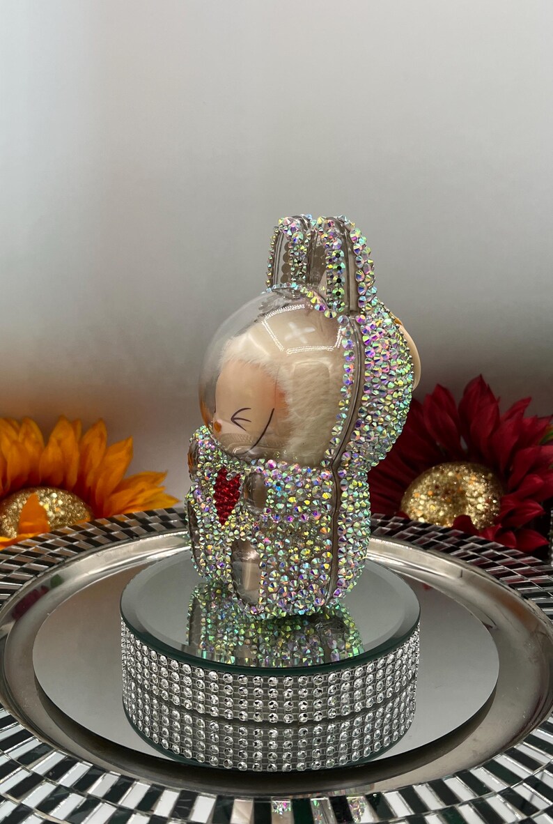 May include: A decorative bunny figurine adorned with iridescent rhinestones. The bunny sits on a mirrored surface atop a rhinestone-studded base. The bunny has a red heart and is surrounded by sunflowers and a red flower.