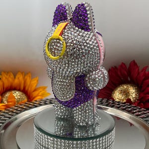 May include: A bedazzled bunny figurine with purple ears and accents, covered in sparkling rhinestones. The bunny has a yellow ring with the text "THE MO" and is displayed on a mirrored platform with a rhinestone border. Decorative flowers surround the figurine.