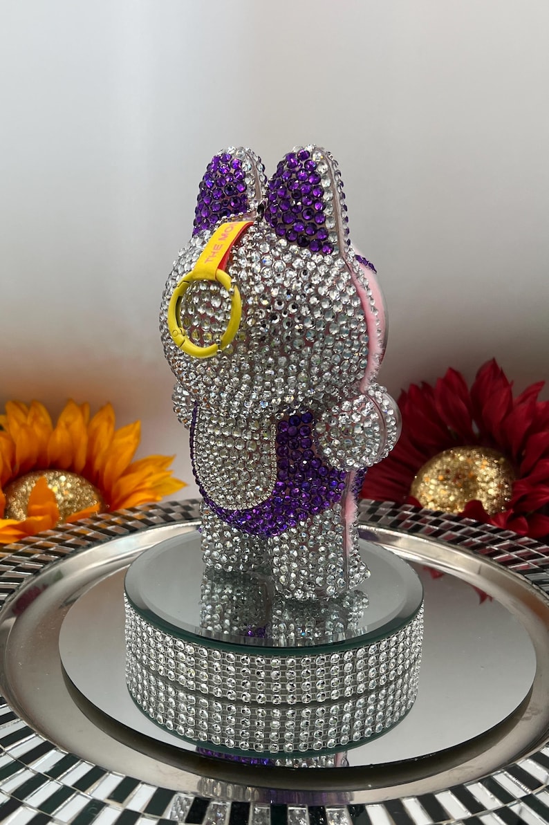 May include: A bedazzled bunny figurine with purple ears and accents, covered in sparkling rhinestones. The bunny has a yellow ring with the text "THE MO" and is displayed on a mirrored platform with a rhinestone border. Decorative flowers surround the figurine.