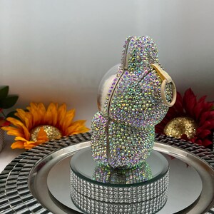 May include: An iridescent clutch bag, encrusted with small, colourful rhinestones. The bag has a rounded shape with a gold-coloured handle and is displayed on a mirrored platform with a rhinestone border. The background includes sunflowers and a decorative tray.