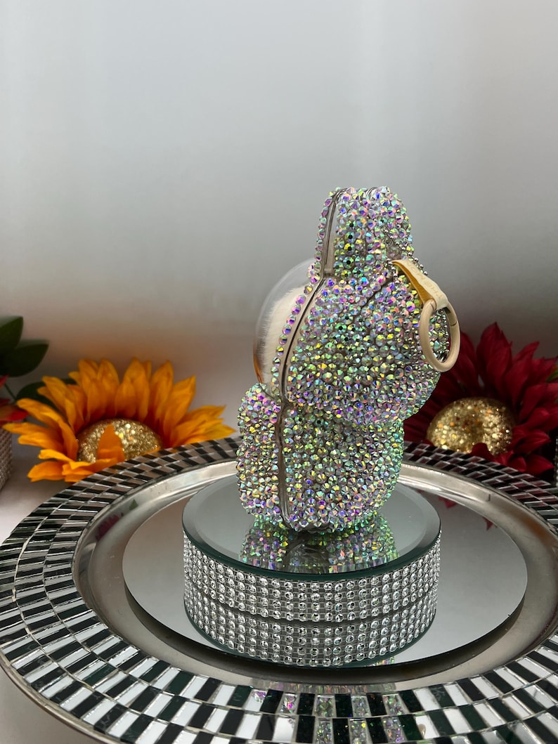 May include: An iridescent clutch bag, encrusted with small, colourful rhinestones. The bag has a rounded shape with a gold-coloured handle and is displayed on a mirrored platform with a rhinestone border. The background includes sunflowers and a decorative tray.
