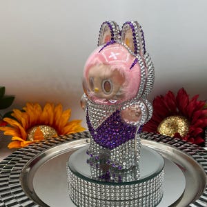 May include: A rhinestone-encrusted figurine of a character in a bunny-eared helmet. The figure has a pink face and a purple and silver outfit. It is displayed on a mirrored platform with a silver tray, with sunflowers in the background.