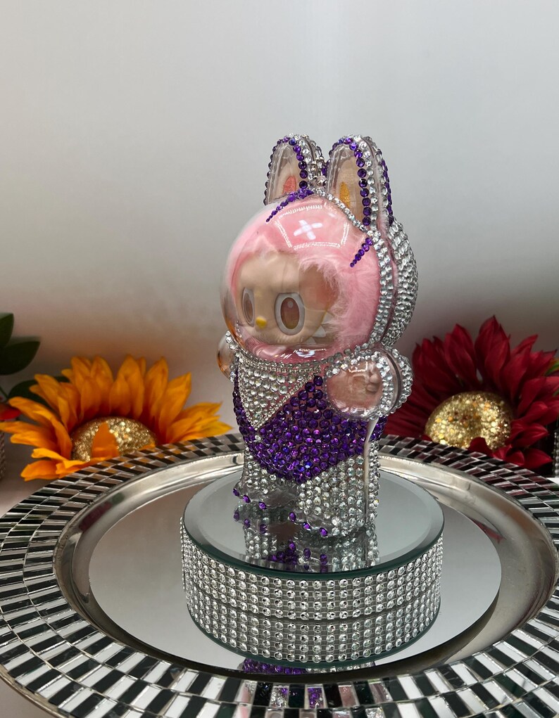 May include: A rhinestone-encrusted figurine of a character in a bunny-eared helmet. The figure has a pink face and a purple and silver outfit. It is displayed on a mirrored platform with a silver tray, with sunflowers in the background.