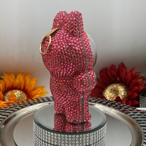 May include: A pink crystal-covered bear-shaped decorative object with a gold-coloured ring. The bear is displayed on a mirrored platform with a silver tray and a decorative, crystal-studded base. The background includes sunflowers.