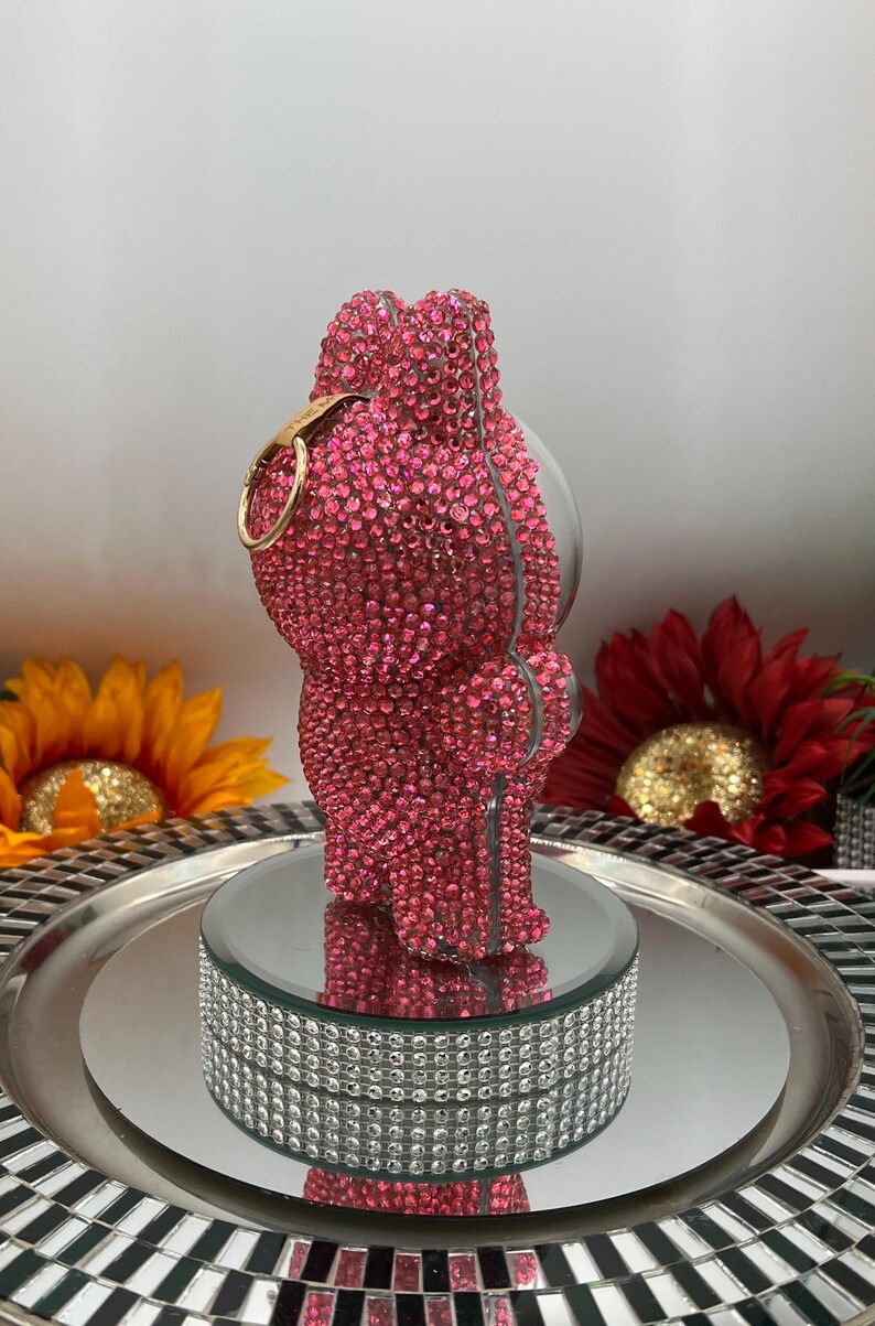 May include: A pink crystal-covered bear-shaped decorative object with a gold-coloured ring. The bear is displayed on a mirrored platform with a silver tray and a decorative, crystal-studded base. The background includes sunflowers.