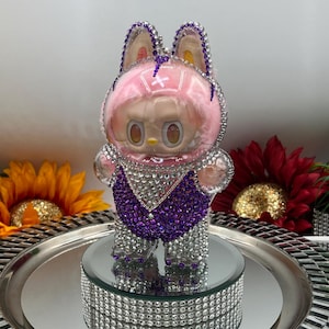 May include: A figurine of a cartoon character, adorned with crystals, featuring a pink fluffy head covering and purple and silver details. It stands on a mirrored surface and a crystal-studded base. The background includes sunflowers.