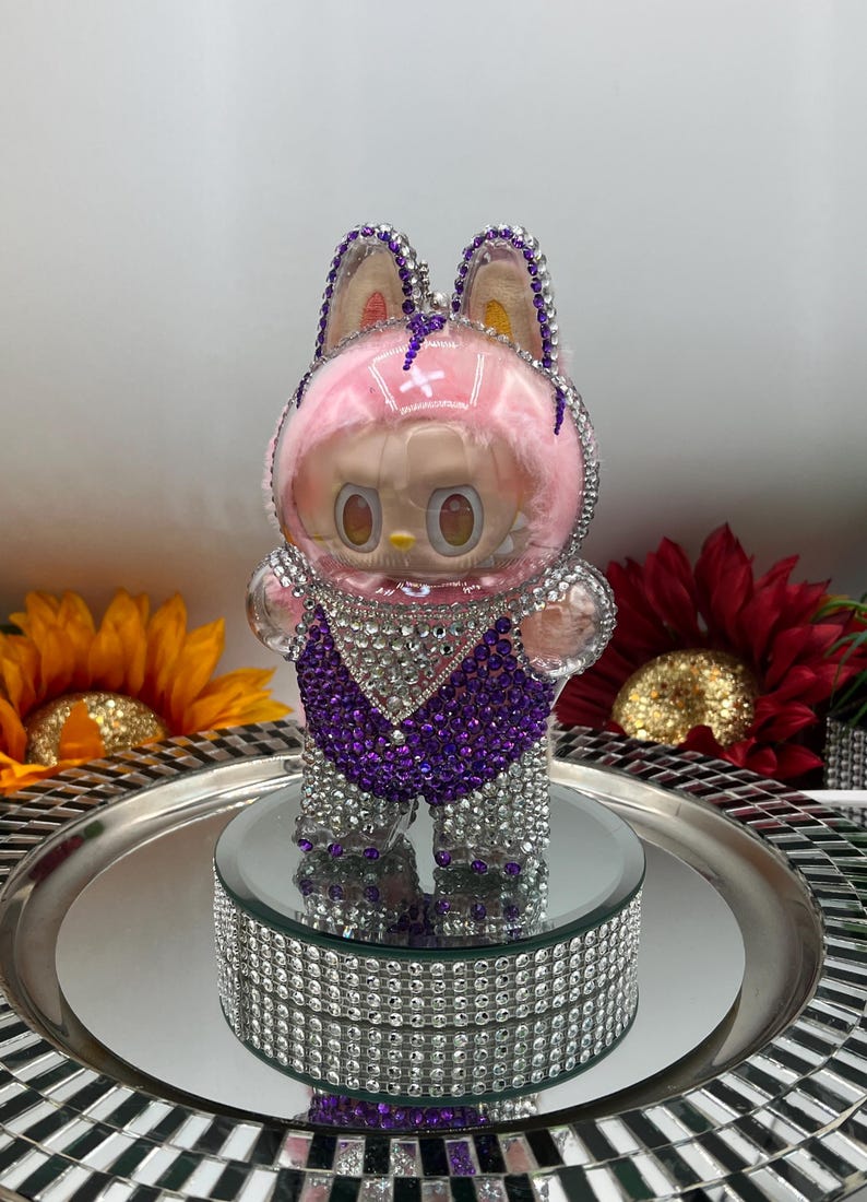 May include: A figurine of a cartoon character, adorned with crystals, featuring a pink fluffy head covering and purple and silver details. It stands on a mirrored surface and a crystal-studded base. The background includes sunflowers.