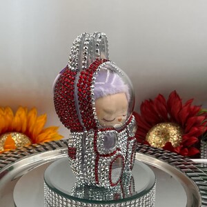 May include: A decorative figurine of a sleeping baby in a red and silver astronaut suit. The suit is encrusted with rhinestones. The figurine sits on a mirrored surface with a rhinestone-covered base, with sunflowers and red flowers in the background.