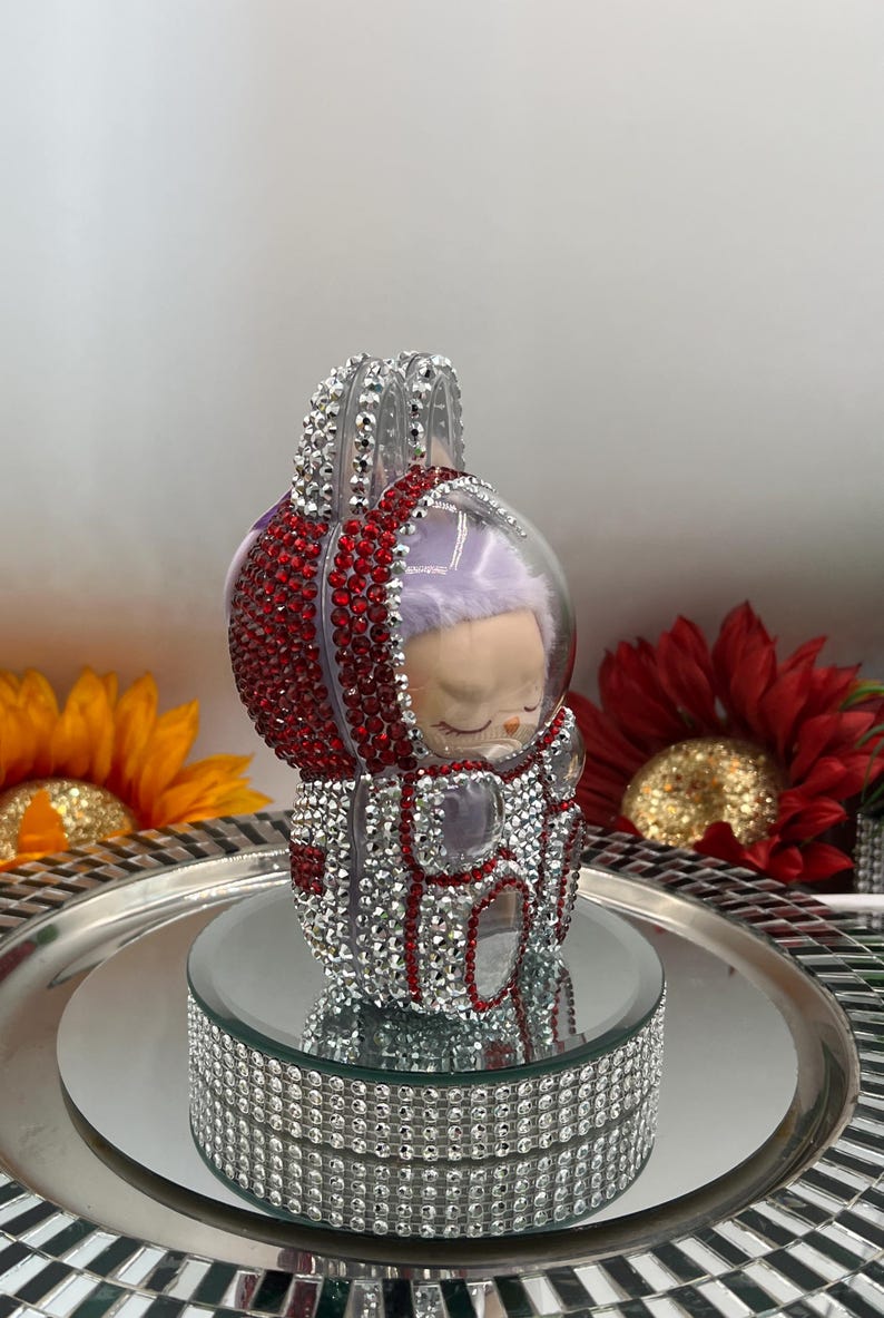 May include: A decorative figurine of a sleeping baby in a red and silver astronaut suit. The suit is encrusted with rhinestones. The figurine sits on a mirrored surface with a rhinestone-covered base, with sunflowers and red flowers in the background.