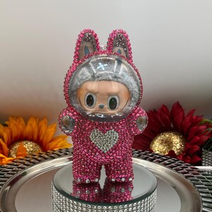 May include: A pink, crystal-covered figurine in a bunny suit, featuring a heart-shaped detail. The figure has a clear helmet and is displayed on a mirrored surface with a decorative, crystal-studded base. The background includes sunflowers and a red flower.