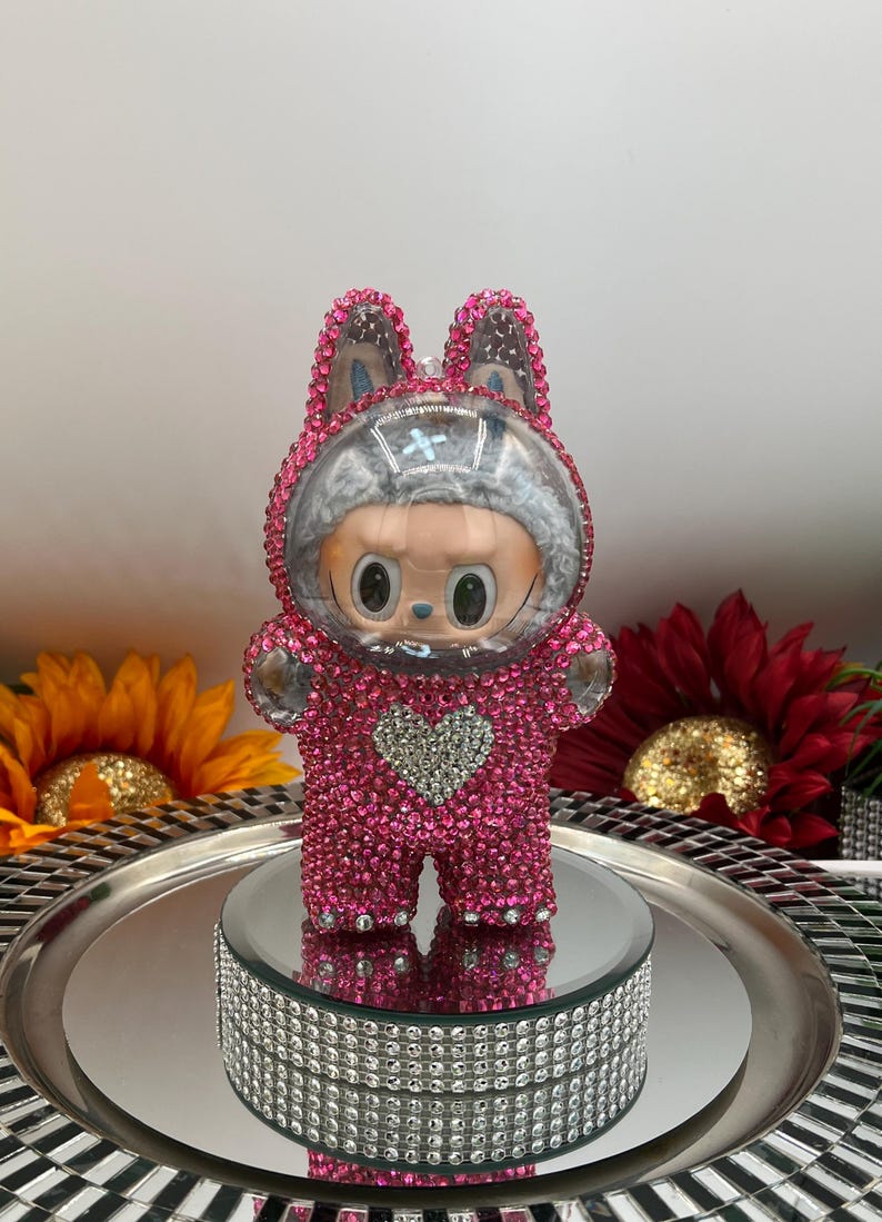 May include: A pink, crystal-covered figurine in a bunny suit, featuring a heart-shaped detail. The figure has a clear helmet and is displayed on a mirrored surface with a decorative, crystal-studded base. The background includes sunflowers and a red flower.