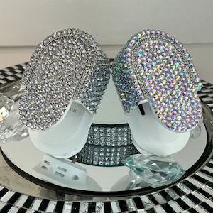 May include: Two white outlet covers with a decorative design of iridescent rhinestones. The outlet covers are on a mirrored surface with a black and white checkered pattern.