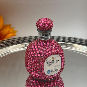 May include: A bedazzled Don Julio Blanco tequila bottle. The bottle is covered in pink rhinestones, including the cap. The label is visible, and the bottle is reflected on a silver tray.