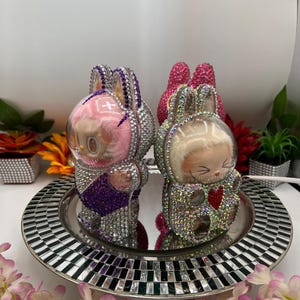 May include: Two rhinestone-encrusted bunny figurines. One is pink and purple, the other silver and gold. They sit on a mirrored tray with a black and white mosaic border, surrounded by flowers. A decorative item.