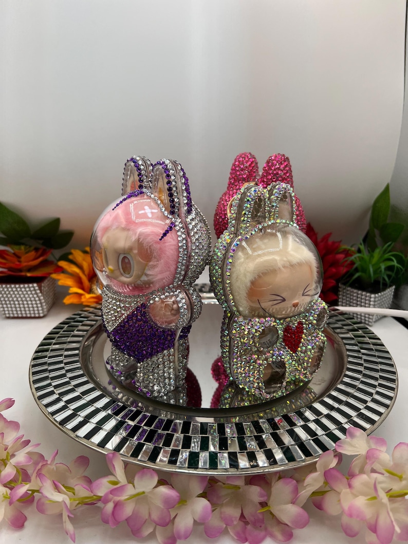 May include: Two rhinestone-encrusted bunny figurines. One is pink and purple, the other silver and gold. They sit on a mirrored tray with a black and white mosaic border, surrounded by flowers. A decorative item.
