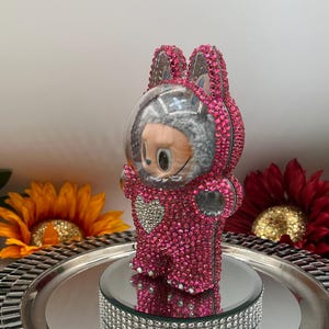May include: A pink, bedazzled figurine of a bunny in a spacesuit. The figurine is covered in small, pink rhinestones and has a clear helmet. It stands on a mirrored platform with a rhinestone border. Decorative sunflowers and a red flower are in the background.