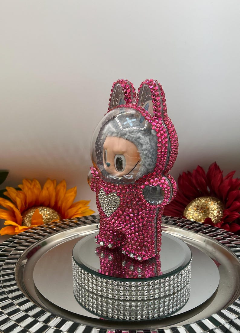 May include: A pink, bedazzled figurine of a bunny in a spacesuit. The figurine is covered in small, pink rhinestones and has a clear helmet. It stands on a mirrored platform with a rhinestone border. Decorative sunflowers and a red flower are in the background.