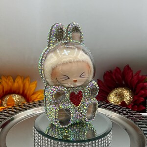 May include: A decorative bunny figurine with a clear, domed head and rhinestone embellishments. The bunny has a red heart on its chest and is displayed on a mirrored tray with a rhinestone border. The background includes sunflowers.