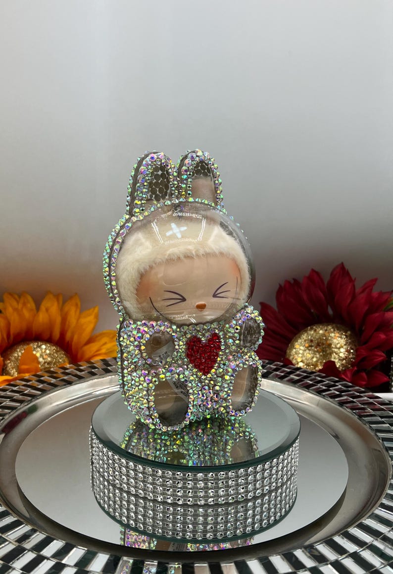May include: A decorative bunny figurine with a clear, domed head and rhinestone embellishments. The bunny has a red heart on its chest and is displayed on a mirrored tray with a rhinestone border. The background includes sunflowers.
