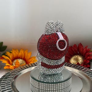 May include: A bunny-shaped ornament, encrusted with red and silver rhinestones. The bunny features silver rhinestone ears and a white loop with text. It rests on a mirrored platform, presented on a silver tray.