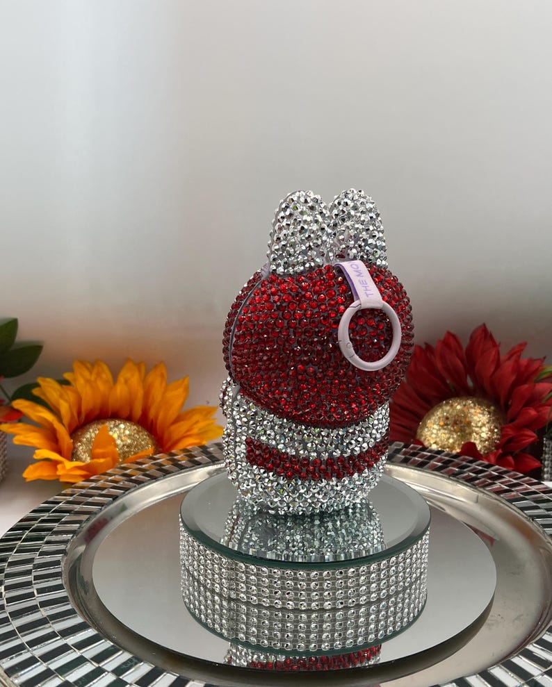 May include: A bunny-shaped ornament, encrusted with red and silver rhinestones. The bunny features silver rhinestone ears and a white loop with text. It rests on a mirrored platform, presented on a silver tray.