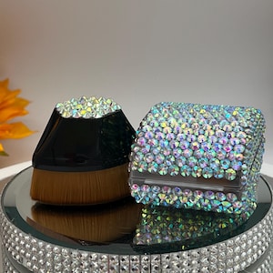 May include: Two makeup brushes with iridescent crystal embellishments. One brush has a black handle and a flat, angled brush head. The other is a rectangular brush holder covered in crystals. Both sit on a mirrored surface.