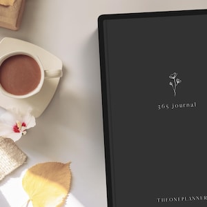 May include: A flat lay featuring a dark gray journal with the text "365 journal" and a floral design. A white teacup with a brown beverage sits on a white saucer. A beige sweater, a yellow leaf, and a white flower are also present.