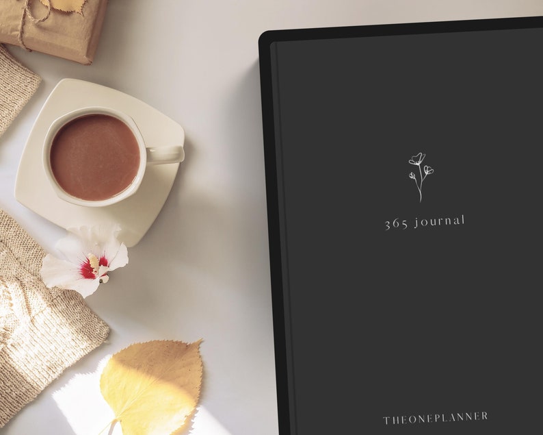 May include: A flat lay featuring a dark gray journal with the text "365 journal" and a floral design. A white teacup with a brown beverage sits on a white saucer. A beige sweater, a yellow leaf, and a white flower are also present.