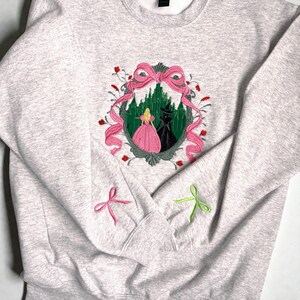 May include: A light gray sweatshirt with an embroidered design featuring a pink bow, a green forest, and two figures. The sleeves have embroidered pink and green bows. The design is centered on the chest.