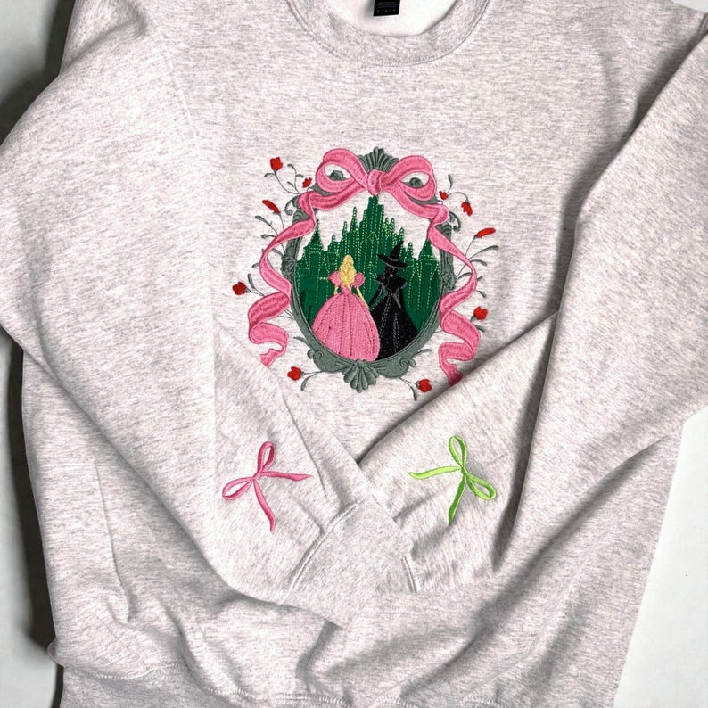 May include: A light gray sweatshirt with an embroidered design featuring a pink bow, a green forest, and two figures. The sleeves have embroidered pink and green bows. The design is centered on the chest.