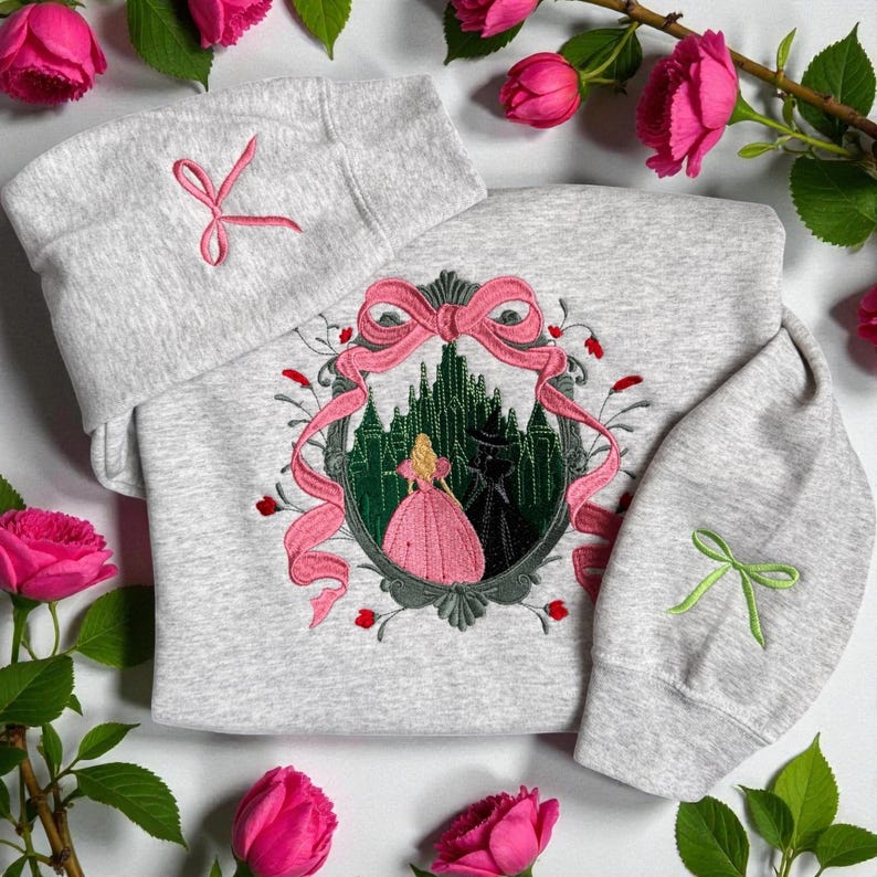 May include: A light gray sweatshirt and matching hat set. The sweatshirt features an embroidered design of a princess in a pink dress in front of a green castle. Pink bows and red roses are also embroidered on the sweatshirt. A pink bow is embroidered on the hat.