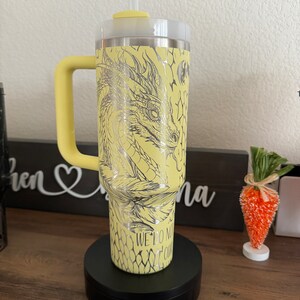May include: Yellow stainless steel tumbler with a dragon design and the text "We do not follow" on the side. The tumbler has a black lid and a black handle.