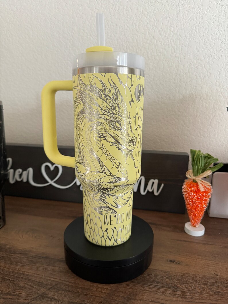 May include: Yellow stainless steel tumbler with a dragon design and the text "We do not follow" on the side. The tumbler has a black lid and a black handle.