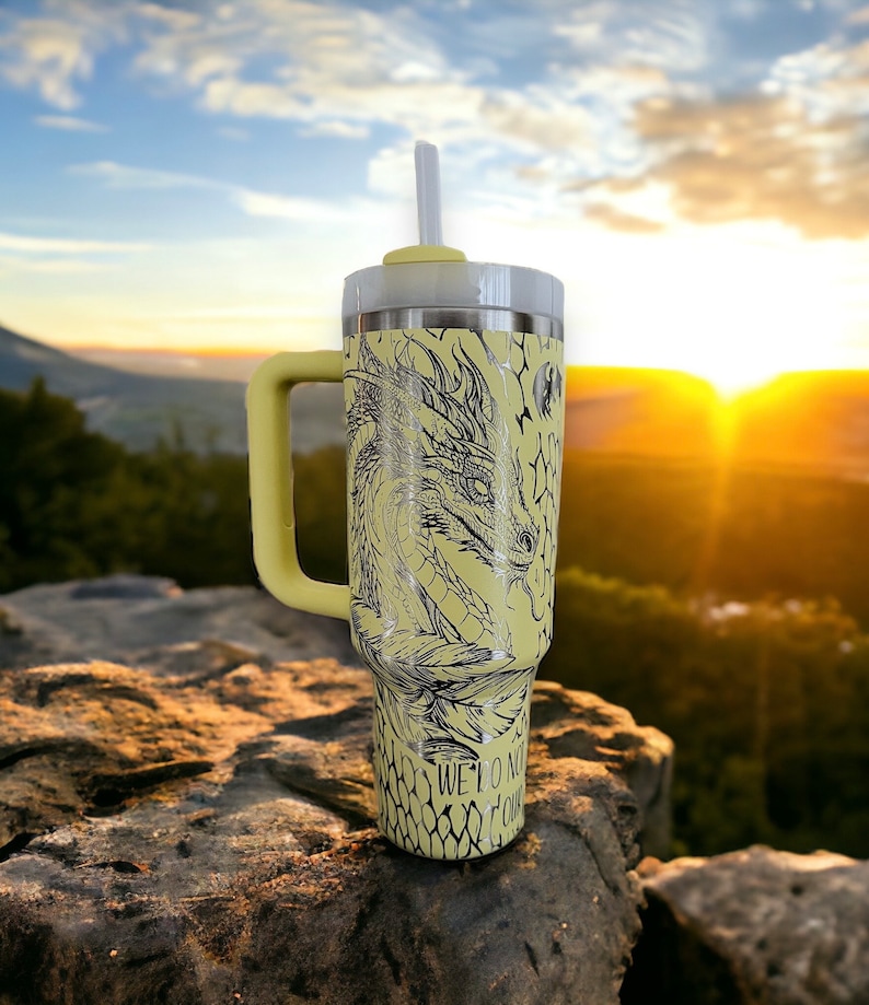 May include: A yellow stainless steel travel mug with a black dragon design and the text "We do not bow to our fears". The mug has a handle and a straw.