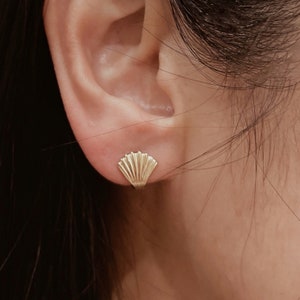 May include: A gold stud earring shaped like a fan or seashell with vertical ridges. The earring is small and metallic, featuring a polished finish that reflects light. It is secured to the earlobe, displaying a simple, symmetrical design.