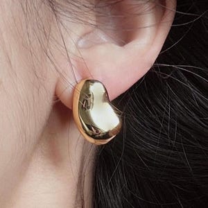 May include: A polished gold-tone earring with a smooth, rounded, and slightly irregular organic shape. The surface is highly reflective, catching light to create bright highlights across its curved, chunky, and solid metallic form.