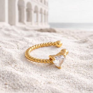 Puede incluir: Un pendiente de oreja dorado con un diseño de cuerda trenzada. El pendiente tiene una gema transparente en forma de corazón. La joya está colocada sobre una playa de arena, con un edificio y el mar al fondo.