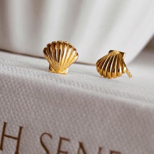 May include: A pair of gold-toned stud earrings shaped like small, ribbed seashells. Each earring features a fan-like design with vertical ridges radiating from the base to the scalloped edge, showcasing a polished, reflective metallic finish.