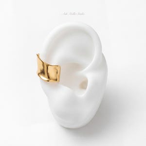 May include: A gold-toned ear cuff with a smooth, undulating, and organic wave-like surface texture. The jewelry piece is displayed on a white ear-shaped model. The text Ash Moltke Studio is visible in small, light grey font above the ear model.