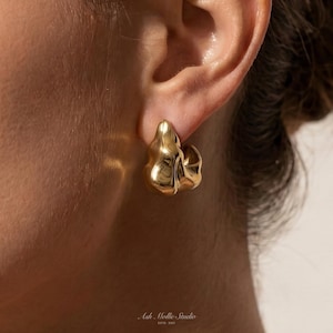May include: A close-up of a gold-coloured earring with an organic, molten design. The earring is a small hoop with a textured surface, reflecting light. The earring is worn on an ear, with the skin in the background.