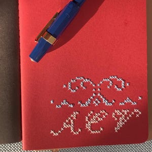 May include: A red notebook with a blue pen resting on top. The notebook has a stitched design in white thread. The pen has a gold-colored clip. The notebook is open, revealing brown pages.