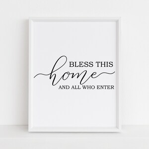 Op de afbeelding: Witte ingelijste print met zwarte kalligrafie met de tekst "Bless this home and all who enter".