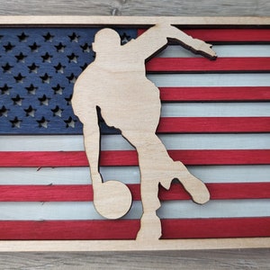 May include: A wooden cutout of a person bowling, superimposed on a red, white, and blue American flag. The flag is made of wood and has a distressed look.