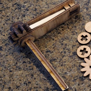 Puede incluir: Un juguete de madera con un brazo largo y delgado y un mecanismo parecido a un engranaje en el extremo. El brazo está unido a una base de madera con una ranura para que una pieza se deslice a través de ella. El juguete está hecho de madera marrón claro y tiene un acabado natural.