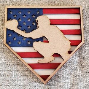 May include: Wooden cutout of a baseball player in a catcher's stance, superimposed on a red, white, and blue American flag. The player is in silhouette, and the flag is in the shape of a baseball diamond.