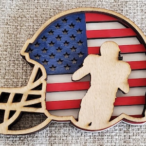 May include: Wooden cutout of a football helmet with an American flag design. The helmet is layered and the flag is cut out to show a silhouette of a football player.