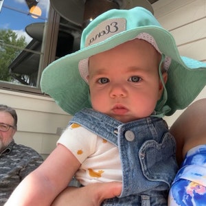 May include: A baby wearing a light blue denim overall and a light green sun hat with the word "angel3" written on it. The baby is being held by a person wearing a floral shirt.