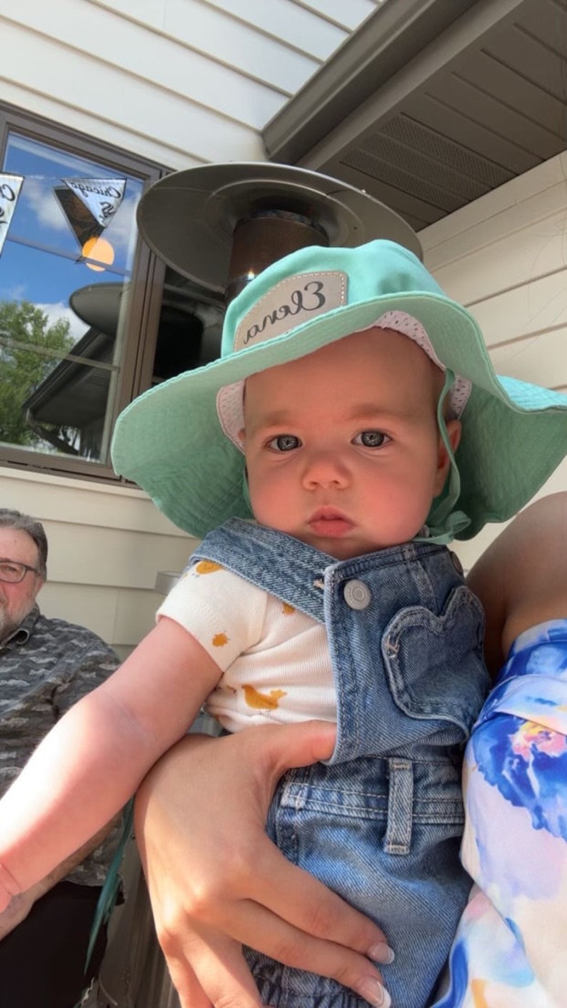 May include: A baby wearing a light blue denim overall and a light green sun hat with the word "angel3" written on it. The baby is being held by a person wearing a floral shirt.
