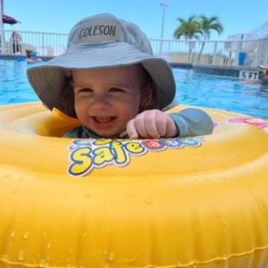 May include: A smiling baby wearing a gray bucket hat with the name "COLESON" embroidered on the front, is sitting in a yellow inflatable pool ring with the word "Safe" printed on it. The baby is in a swimming pool.