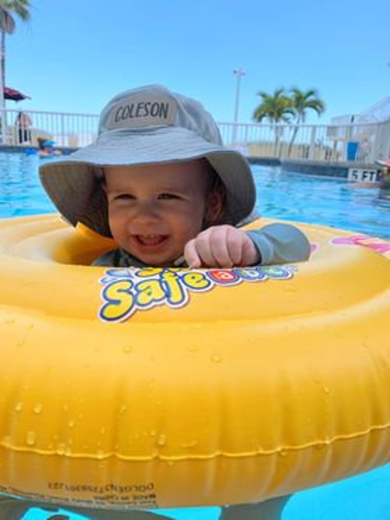 May include: A smiling baby wearing a gray bucket hat with the name "COLESON" embroidered on the front, is sitting in a yellow inflatable pool ring with the word "Safe" printed on it. The baby is in a swimming pool.